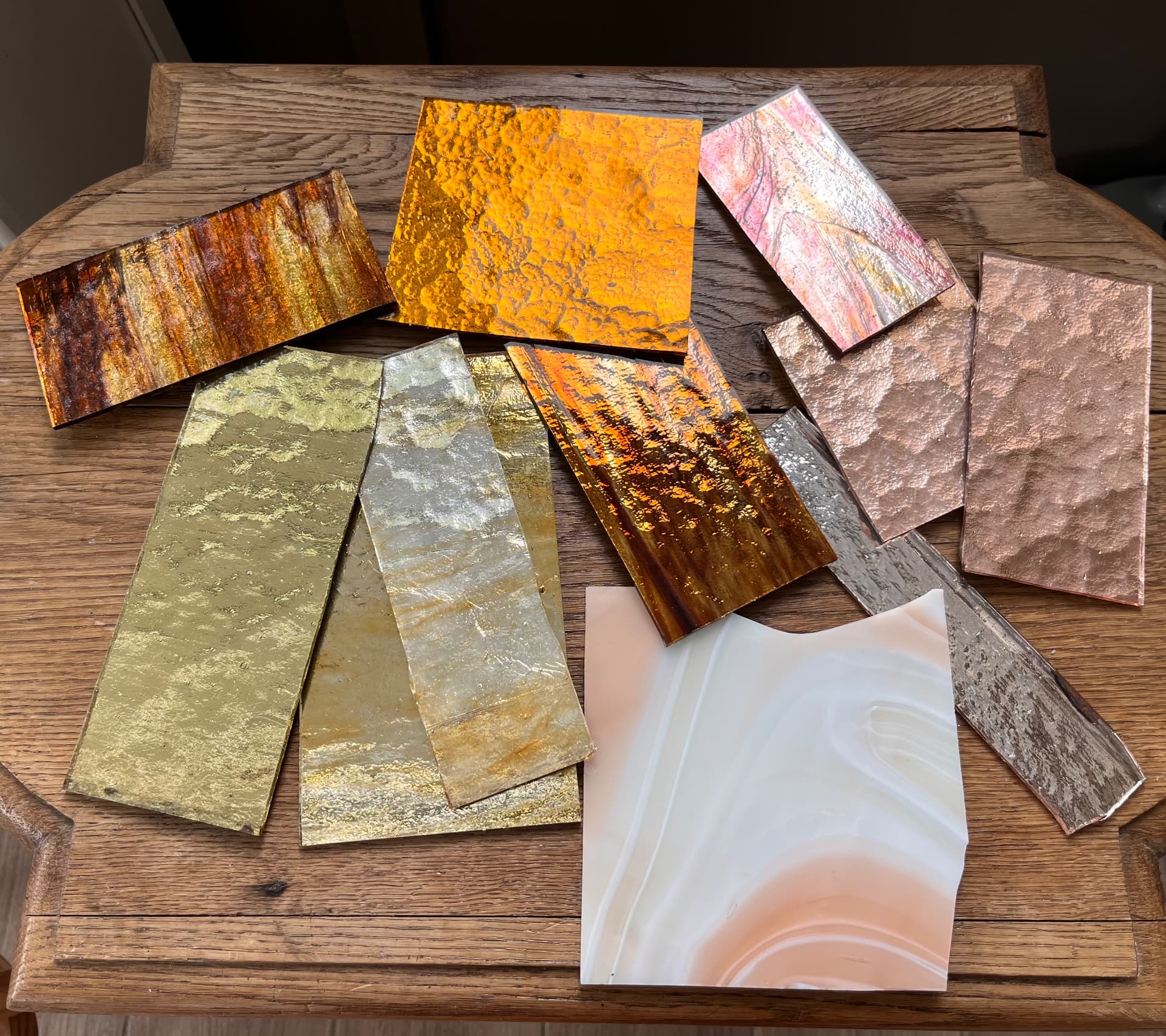 Plaques de verre dorées, orangées, argentées et rosées aux textures variées posées sur une table en bois
