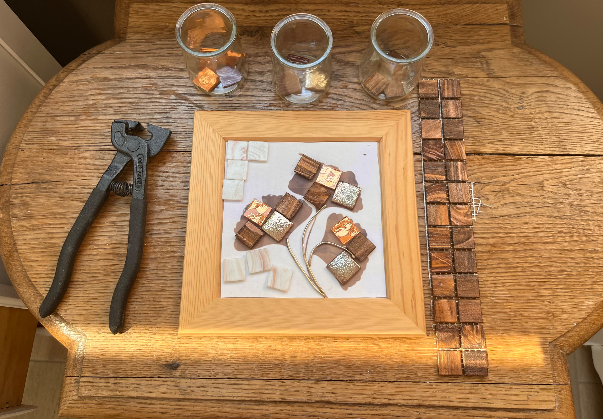 Vue du dessus d’une table en bois avec une pince, un cadre en cours de mosaïque végétale et des tesselles dorées